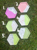 Six hexagonal concrete trays with a terrazzo pattern, painted in pastel pink, purple, green, and white, arranged on a green grass lawn.