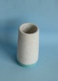 A small cylindrical ceramic vase with a speckled white glaze, pastel pink and blue flecks, and a turquoise base, set against a solid light blue background.